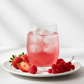 Fotografía realista de un vaso de vidrio con agua de sabor color rojo claro, preparada con jarabe de granadina sin azúcar, con hielo y frutos rojos frescos alrededor. Estilo saludable y gourmet francés, iluminación natural suave, fondo claro minimalista, sin texto ni personas. Formato cuadrado.